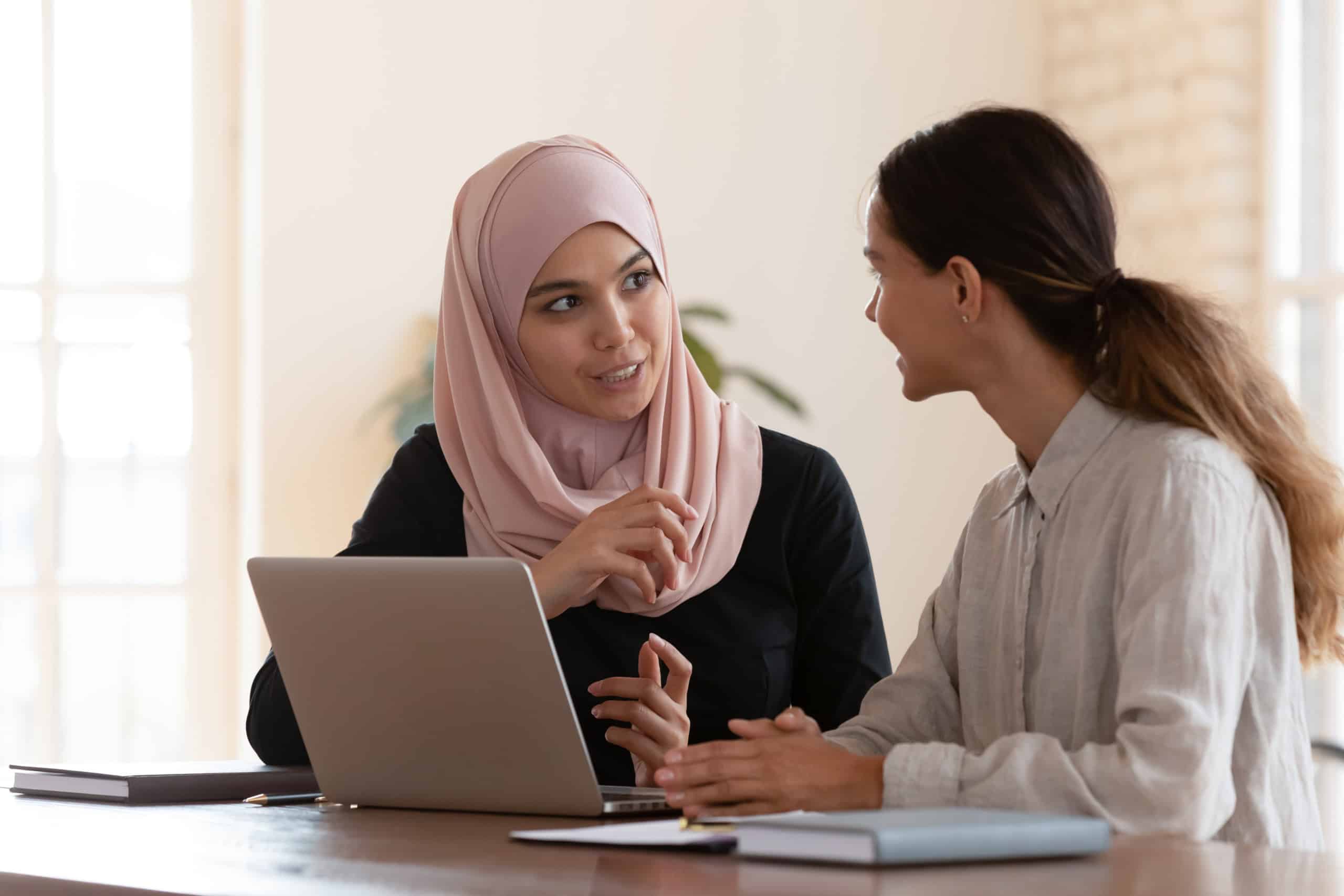 Multiracial young female employees work on laptop discuss business cooperation ideas at meeting together, international businesswomen brainstorm collaborating using computer at office briefing