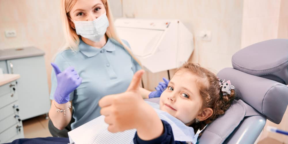 Focus on girl's hand giving thumbs up while woman dentist in medical mask sitting beside kid, showing approval gesture after dental procedure. Concept of pediatric dentistry and dental care approval.