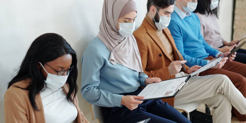 Group of young multi-ethnic job candidates in masks sitting in row and examining resume before interview